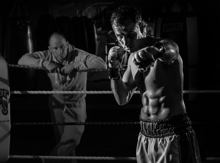 Boxer practicing in the ring with coach observing, captured in dramatic lighting.