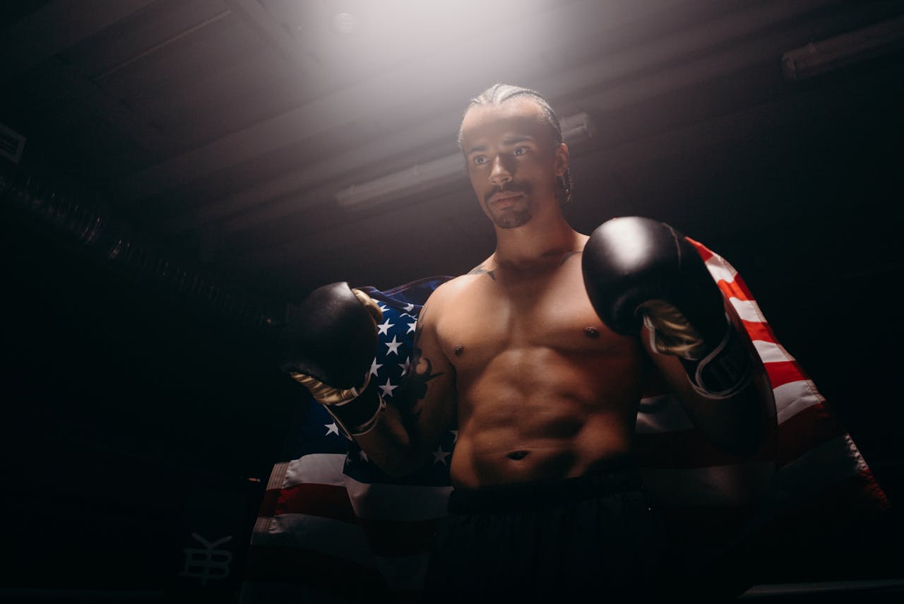 Shirtless boxer with boxing gloves stands confidently with American flag backdrop, embodying strength and patriotism.